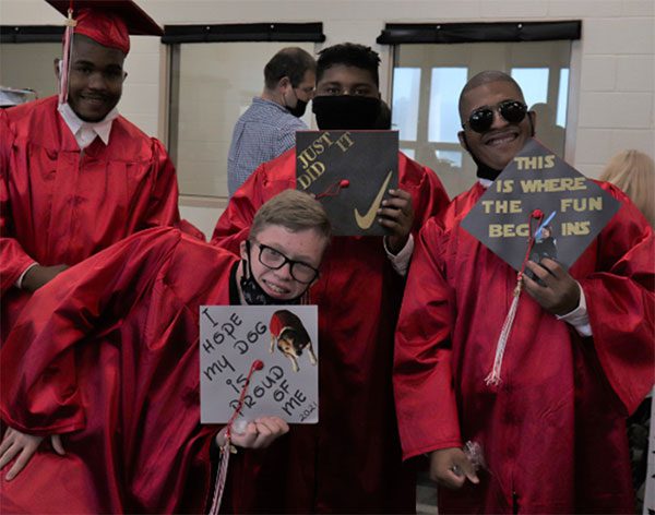 BCSSSD graduates proudly hold up their decorated caps. One says, “I hope my dog is proud of me” and another has the Nike swoosh and says, “Just did it.”