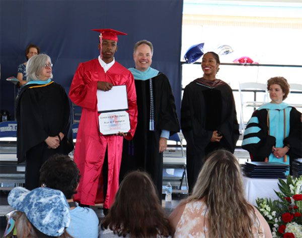 BCSSSD administrators smile behind a graduate on stage, who proudly displays his diploma during the commencement ceremony.