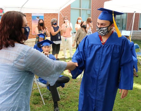 A BCSSSD graduate wearing a traditional cap and gown, along with a facemask, shakes the hand of an administrator following the commencement ceremony.