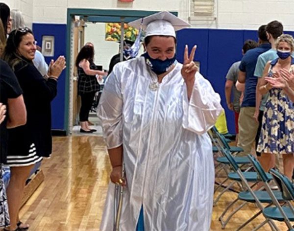 A GCSSSD graduate in a cap and gown puts up the peace sign as she walks during the graduation procession while the crowd claps in the background.