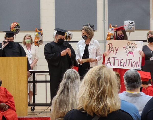 Graduates and administrators at CMCSSSD gather on stage during the commencement ceremony. While one student addresses the crown, another holds up a sign that says “pay attention” to remind those in the audience to be quiet and listen.