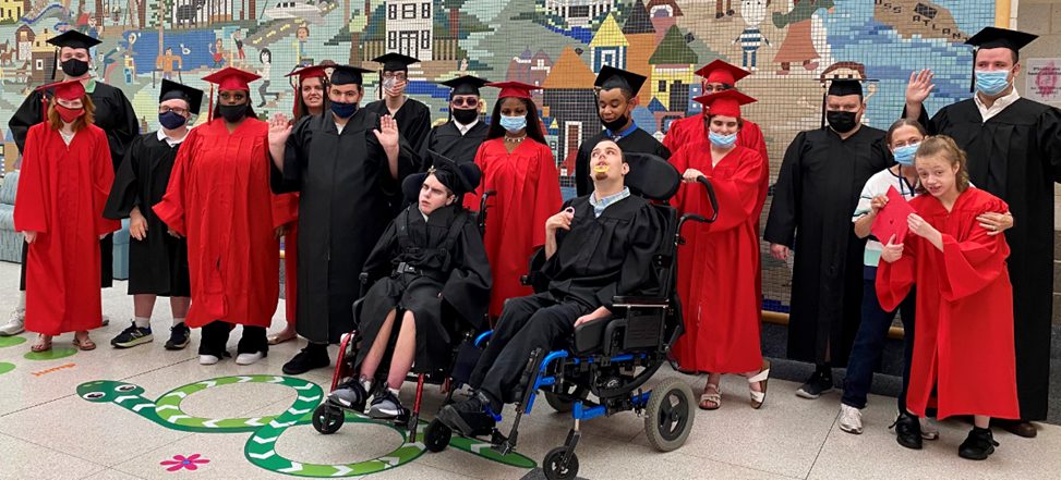 CMCSSSD graduates dressed in red and black caps and gowns gather together for a photo following the commencement ceremony, some are wearing masks, some are waving at the camera.