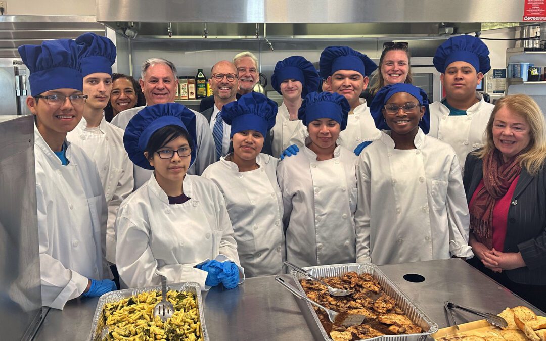 Teenaged students dressed in white chef coats and blue chef hats pose for a photo in Bergen County Special Services School District's commercial kitchen used for culinary instruction. Food they prepared for a special lunch is in pans in front of them.