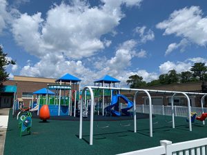 Colorful playground equipment such as swings, slides, a covered chair, talk tubes and play huts are arranged on rubber safety matting and enclosed in an area surrounded by white vinyl fencing.