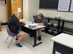 A student is seated at a table sorting dominoes in a box while his teacher watches him from her seat across the table.