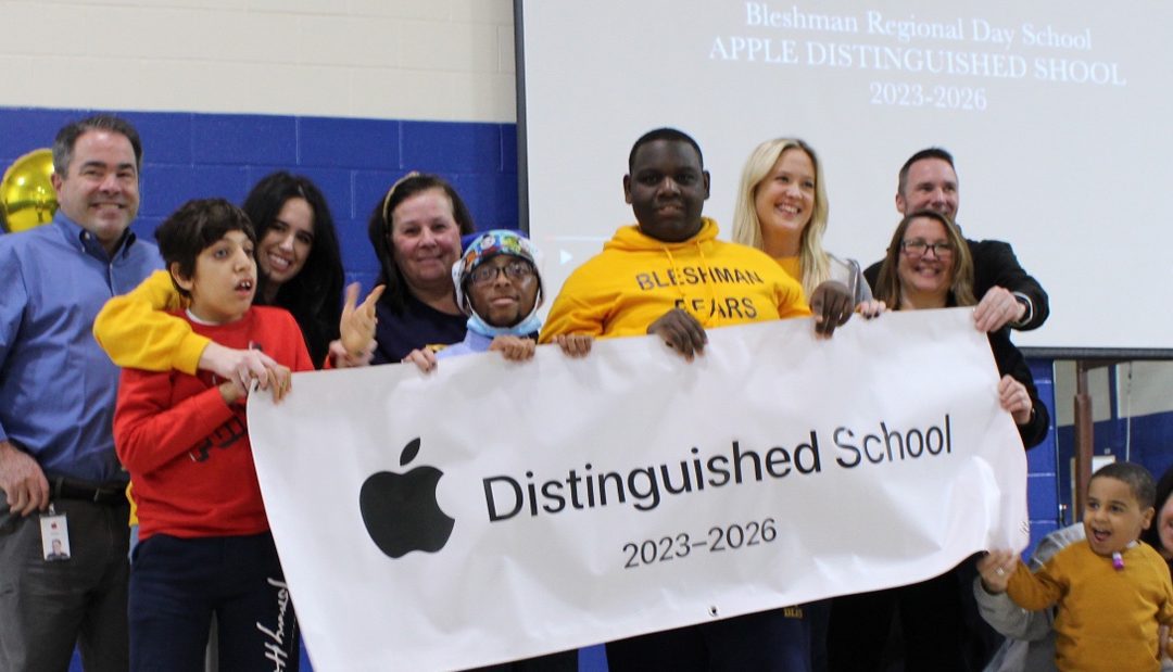 Apple representatives stand on each side of a line of students and school staff members who are holding a banner across the front of the group that says Distinguished School 2023-2026.
