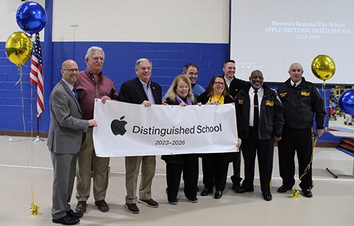 Guests at the Apple Distinguished School recognition ceremony - dressed in suits, dress clothes and uniforms and standing in a line - hold a banner across the front of the group that says Distinguished School 2023-2026.