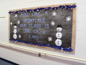 A hallway bulletin board is decorated with a gray wooden background and white snowflakes, snowmen with black buttons and a black top hat, and blue garland. It reads just like a snowflake our class is one of a kind.