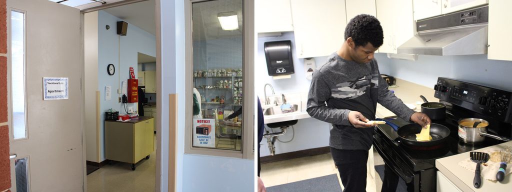 An open classroom door has a laminated white paper sign with a blue decorative border that reads Vocational Lab Apartment on it. A student in a gray shirt is adding a slice of cheese to the grilled cheese sandwich that he’s cooking in a pan on the stovetop. 