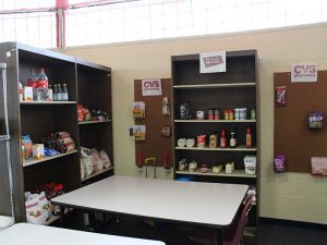 A workstation set up to simulate CVS and Shop Rite store shelves is stocked with products and hanging items. A table with a chair sits in front of the shelves.