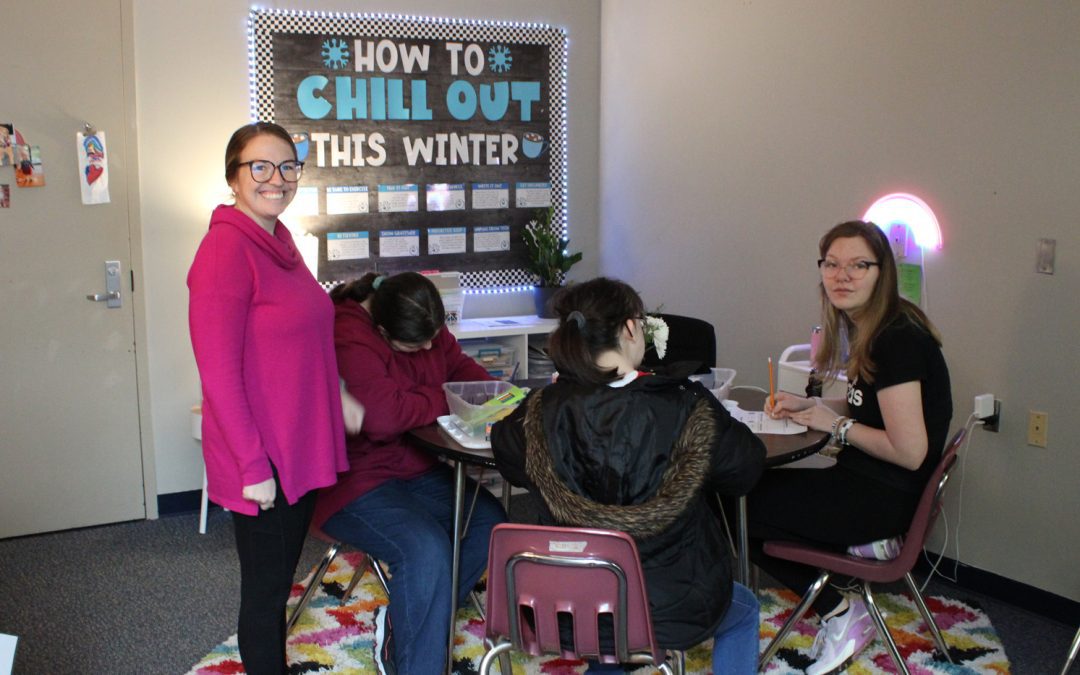 Three female students are seated at a round table with a female teacher standing beside them in a room with dimmed lighting and a how to chill out this winter bulletin board in the background. The table is sitting on a square rug with a rainbow zig-zag pattern.