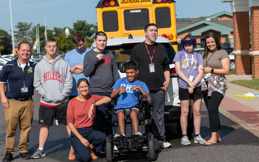 Students are standing outside in a parking lot at the rear of a school bus outside of a school.