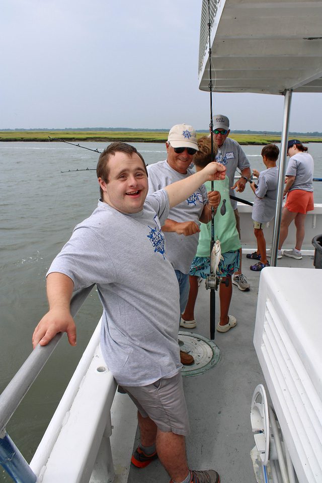 A participant is standing against the side of the boat while holding his fishing line that has a fish on the end of it.