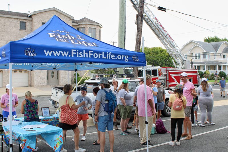 A blue Fish for Life tent has people gathered under it to register while a fire truck is in the background.