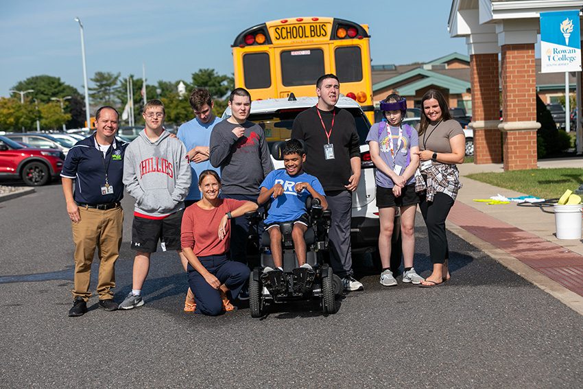 Students are standing outside in a parking lot at the rear of a school bus outside of a school.