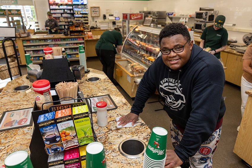 A young man smiles as he cleans the counter of a coffee station at a local cafe.