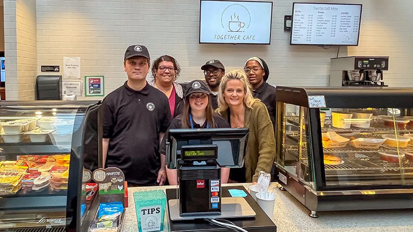 GCSSSD students and staff members stand at the cash register behind the counter of a cafe, with glass food cases on either side of them.