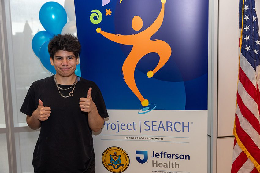 A student is standing with two thumbs up next to a Project SEARCH banner.