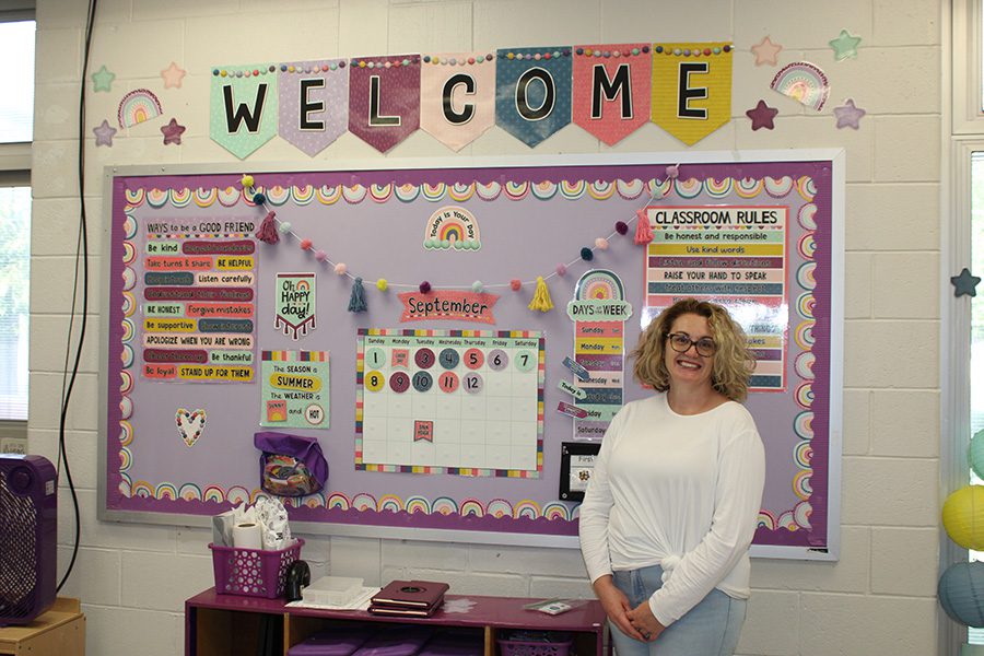 A female teacher stands with her hands folded in front of a colorful bulletin board decorated with rainbows and purple border.