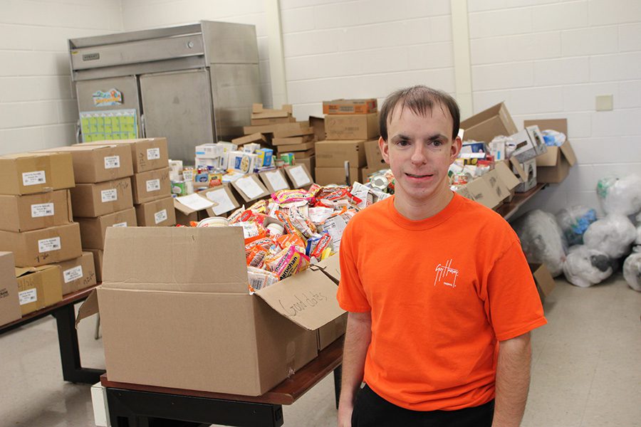 A smiling young man in an orange shirt stands in front of boxes full of food items in the school’s food pantry.