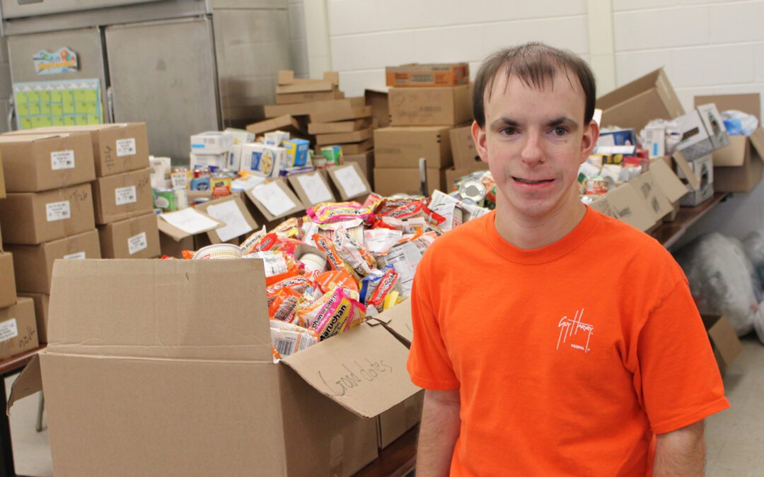 A young man in an orange shirt stands in front of tables full of boxed food items in a food pantry.