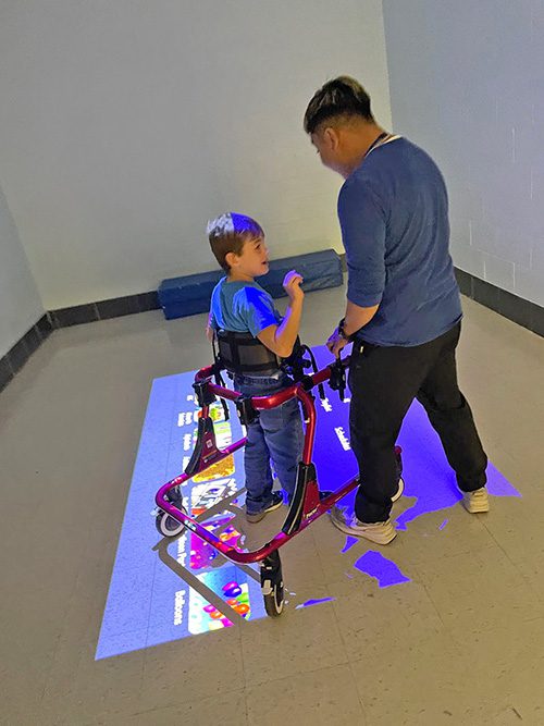 A student in an assistive standing device is working with a teacher to play a game using his feet to control a touch image projected onto the floor.