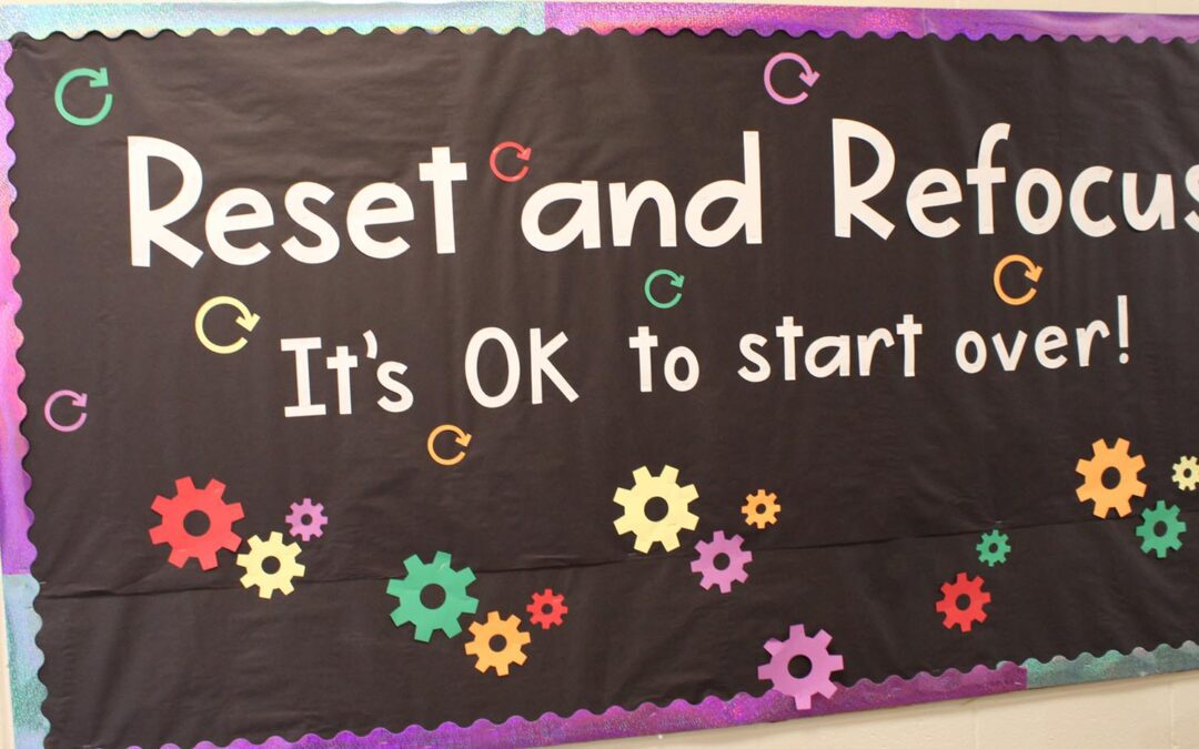 A black bulletin board with white letters and multi-colored gear-shaped cutouts says “Reset and Refocus: It’s OK to start over.”