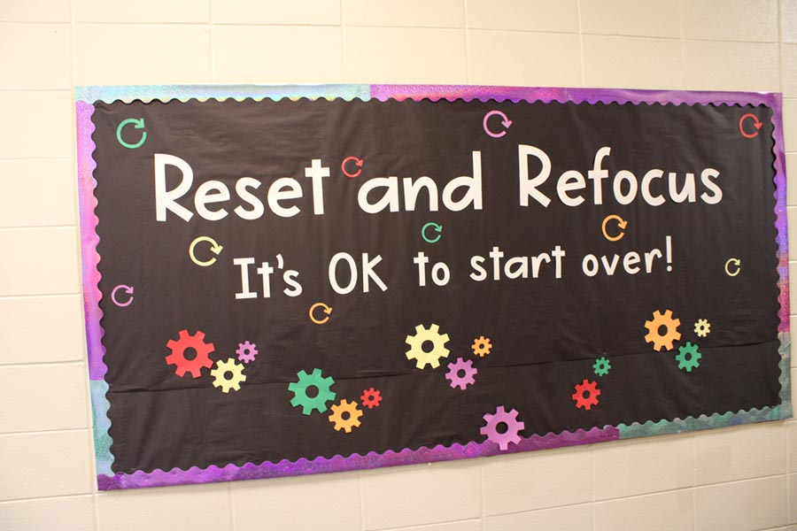 A black bulletin board with white letters and multi-colored gear-shaped cutouts says “Reset and Refocus: It’s OK to start over.”