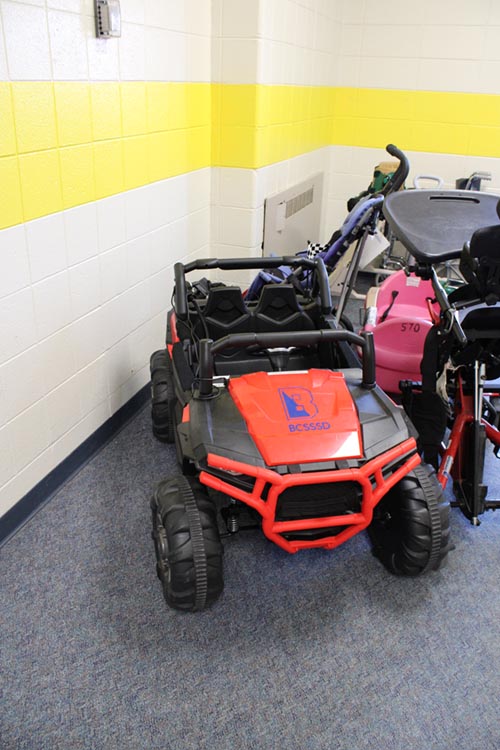 A mini four-wheel utility vehicle with red paint and a BCSSSD logo on the hood sits in the hallway next to other mobility aids.