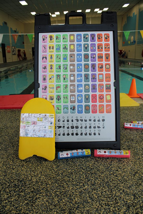 A stand-up sign, pool kick board, and pool noodles, all with communication boards attached to them, are set up on the pool deck.