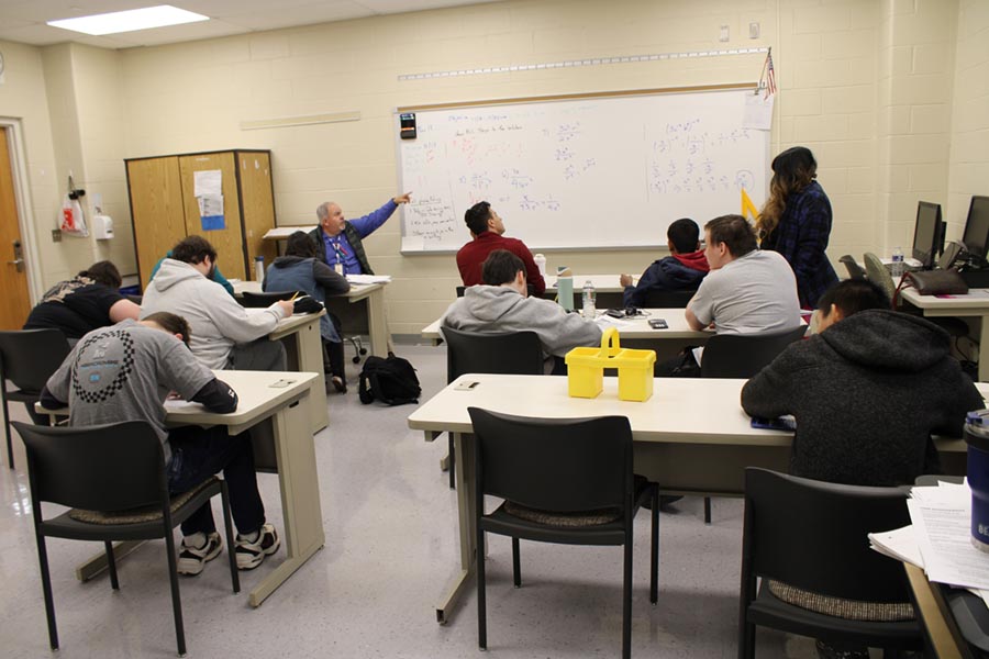 Students are arranged in a traditional classroom, facing the teacher as he points to the lesson on the white board at the front of the room.