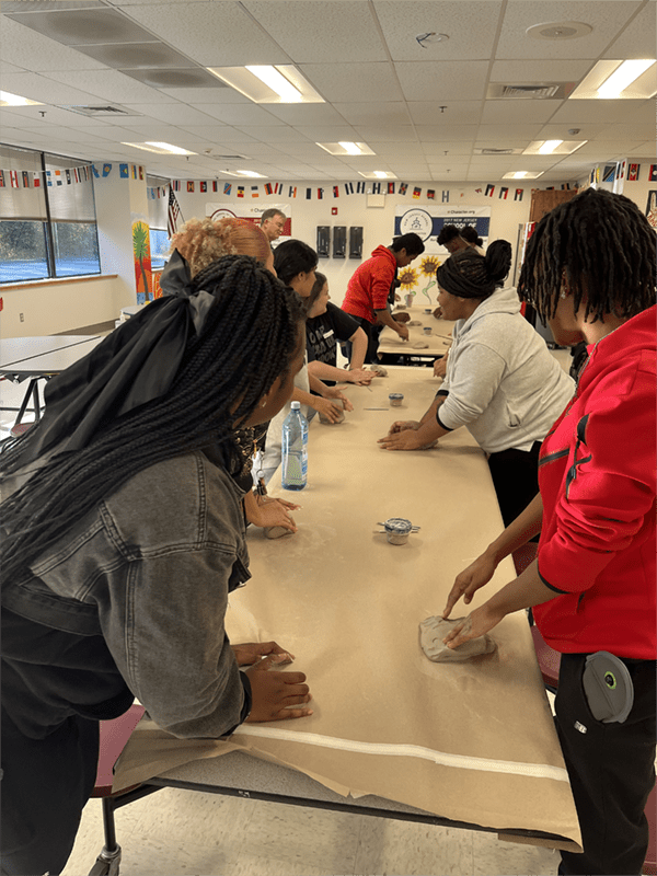 Students are standing along both sides of a long table covered in craft paper; each one is kneading a ball of clay.