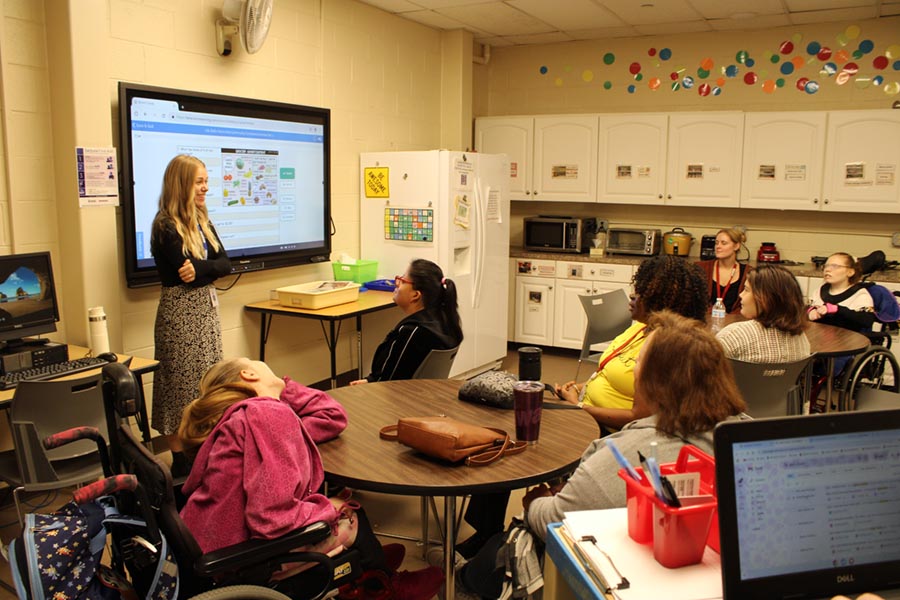 A related service provider stands in front of a classroom of students who are seated in small groups at round tables.