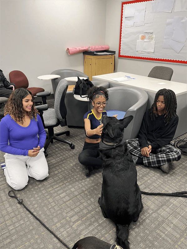 Three students are sitting on the floor of a room with a black dog in the middle; one student is reaching out to pet the dog.